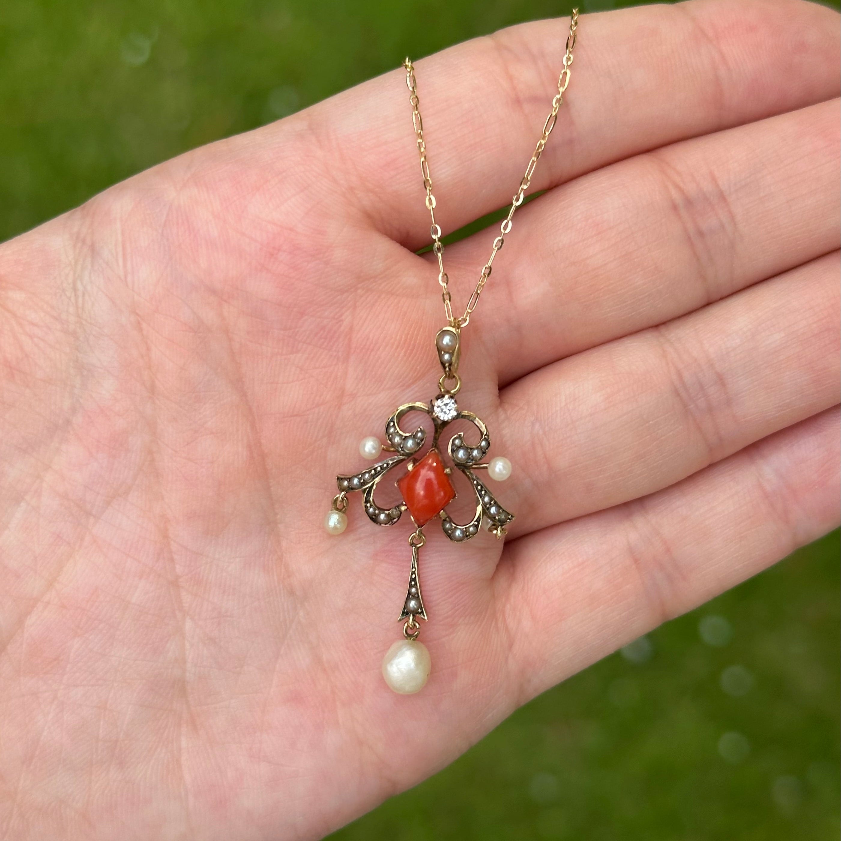 A delicate gold chain necklace with a vintage-style filigree pendant, featuring a central orange-red coral bead, small white pearls, and clear rhinestones, resting on an open hand against a blurred green background.