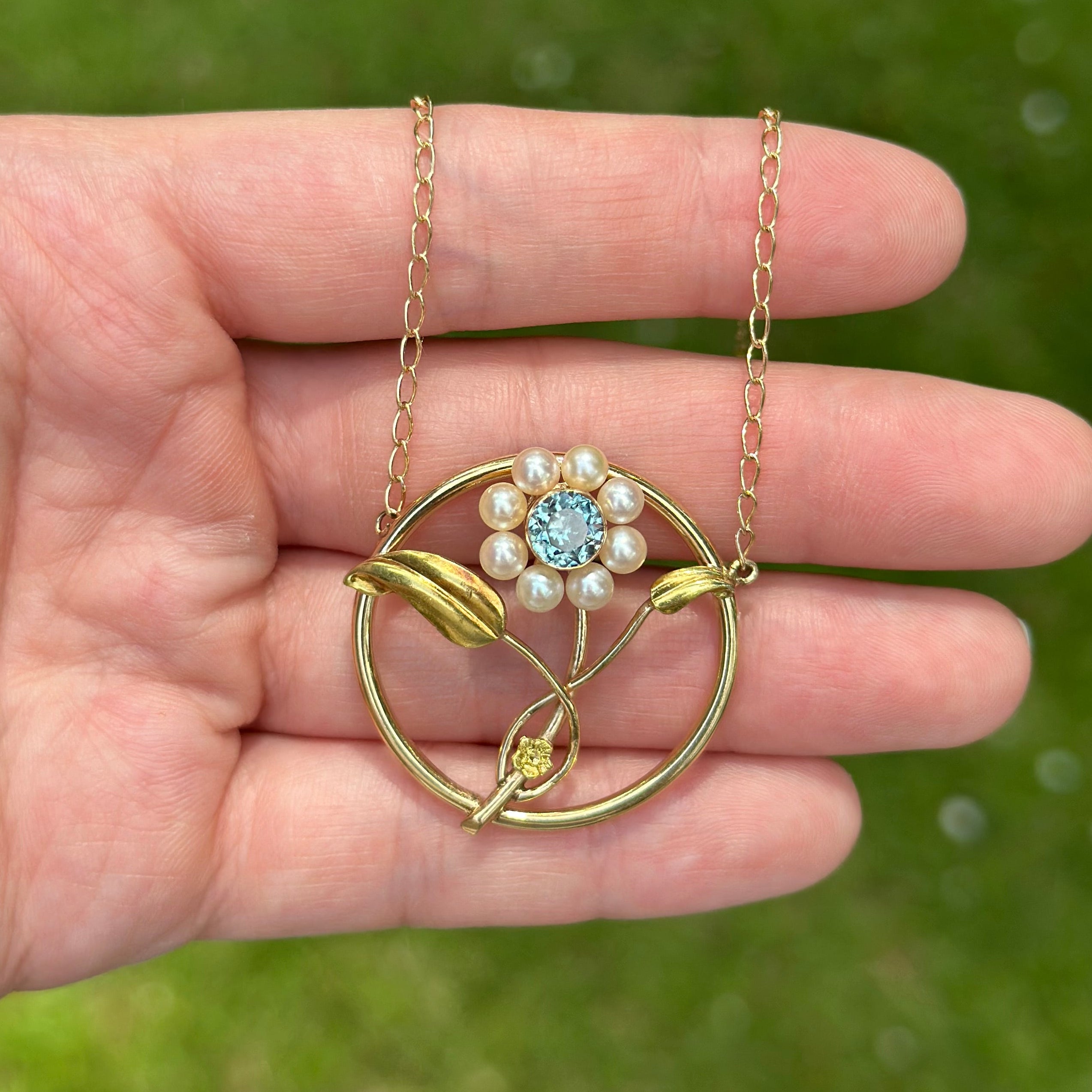 A gold pendant necklace with a circular openwork design, leaf details, and a central floral cluster of white pearls and a light blue gemstone, held in a person's hand against a blurred green grass background.